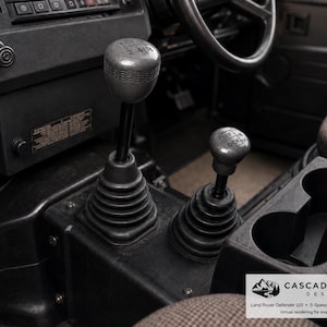 May include: A Land Rover Defender 110 interior featuring a 5-speed gear shift knob and a transfer case knob, both with black rubber gaiters. The gear knob displays a 1-5 gear pattern, while the transfer case knob shows Diff Lock H-N-L settings.