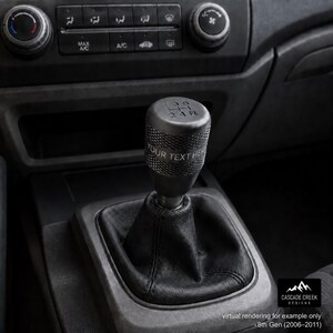 May include: A black cylindrical manual gear shift knob with a knurled texture and a gear pattern on top showing 1, 3, 5, 2, 4, and R. The side of the knob features the engraved text YOUR TEXT HERE, and it is attached to a black leather shift boot.