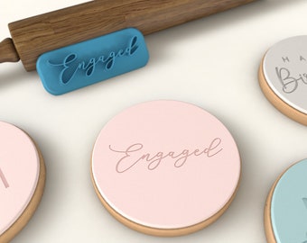 May include: A blue rectangular cookie stamp with the word "Engaged" in cursive script sits next to several round cookies. One cookie is stamped with "Engaged" and another with "Happy Birthday". The cookies have a pink or light blue frosting.