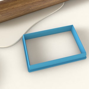 May include: A blue plastic rectangle cookie cutter on a white surface.