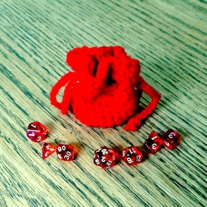 Może przedstawiać: Czerwony, dziany woreczek ze sznurkiem do ściągania z sześcioma czerwonymi kostkami do gry na drewnianej powierzchni.