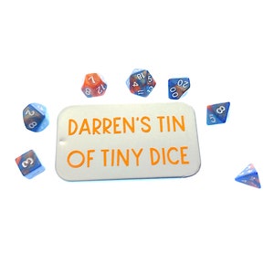 May include: A white metal tin with the text "Darren's Tin of Tiny Dice" in orange lettering.  The tin is surrounded by eight small blue and orange dice with white numbers.