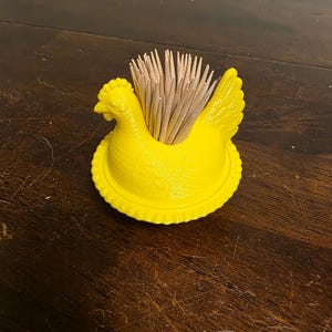 Könnte beinhalten: Ein Set aus drei Keramikbechern mit gesprenkelter, cremefarbener Glasur. Jeder Becher zeigt ein handgemaltes Design: ein blau-grünes Blatt, eine gelbe Blume und eine rote Beere. Die Becher haben eine einfache, abgerundete Form.