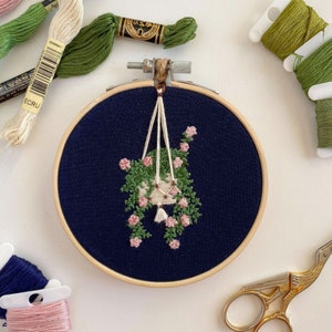May include: An embroidery hoop with navy blue fabric displays a hand-stitched design of a hanging basket with pink flowers and green foliage. Embroidery threads in various colours and scissors are arranged around the hoop.