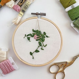 Op de afbeelding: Een witte borduurring met een geborduurd hangend plantontwerp in groen en roze. De ring ligt op een wit oppervlak met borduurgaren en een schaar.