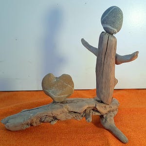 Könnte beinhalten: Eine Skulptur aus Treibholz und Steinen, die eine Figur mit einem runden Steinkopf und einem vertikalen Treibholzkörper mit ausgestreckten Armen darstellt. Ein herzförmiger Stein liegt auf einer Treibholzbasis. Der Text HOME ist sichtbar.