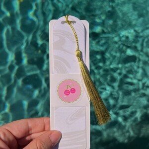 May include: White bookmark with a marble pattern and a gold tassel. The bookmark has a pink and green sticker with the text "happy vibes" and a cherry graphic.