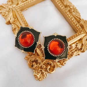 May include: A pair of gold-tone earrings with black enamel and red glass stones. The earrings are in the shape of a square with rounded corners.
