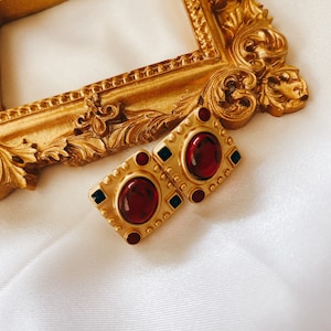 May include: A pair of gold-tone earrings with red and green stones. The earrings are square-shaped and have a textured design.
