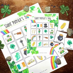 Könnte beinhalten: Ein Saint Patrick's Day Bingo-Spiel mit einem farbenfrohen Regenbogen- und Kleeblatt-Design. Das Spiel beinhaltet eine Bingokarte mit Bildern eines Kobolds, eines Topfes mit Gold, eines vierblättrigen Kleeblatts, einer Harfe, eines Kreuzes, einer Geige, eines Regenbogens, einer Goldmünze, eines Kleeblatts und der Zahl 17. Das Spiel beinhaltet auch einen Satz von Rufkarten mit den gleichen Bildern.