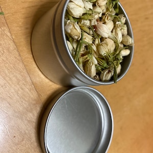 May include: A silver tin filled with dried white jasmine flowers. The tin has a matching lid.