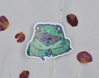 May include: A green frog sticker with a holographic finish. The frog has a wide open mouth and is sitting on a white background.