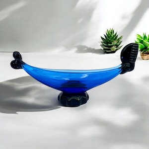 Peut inclure: Un bol en verre bleu cobalt en forme de bateau avec des accents en verre noir. Le bol est sur une surface blanche.