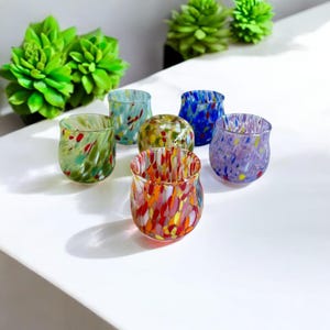 Può includere: Un set di sei bicchieri di vetro maculato dai colori vivaci: verde, blu, giallo, rosso e viola. I bicchieri sono disposti su una superficie bianca, con piante succulente verdi sullo sfondo. Ideali per servire bevande.