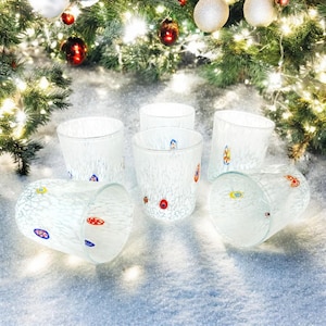 Könnte beinhalten: Sechs klare, strukturierte Glasbecher mit farbenfrohen, dekorativen Akzenten. Die Gläser sind auf einer schneebedeckten Oberfläche angeordnet, mit einem Weihnachtsbaum und Ornamenten im Hintergrund. Die Gläser sind etwa 10 cm hoch.