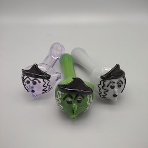 May include: Three glass pipes with witch faces. The pipes are purple, green, and white. Each pipe has a black hat and a white face with black eyes and a nose.