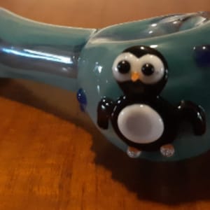 May include: A teal glass pipe with a penguin design. The pipe has a rounded bowl and a long, curved stem. The penguin is black and white with large eyes and is centered on the bowl.