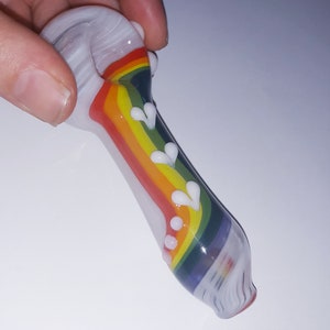 May include: A small glass pipe with a rainbow design and white accents. The pipe is made of clear glass and has a rounded bowl.