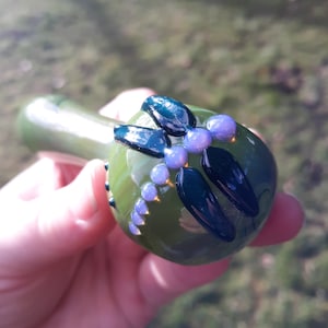 May include: A green glass pipe with a decorative design of dark green leaves and purple dots.