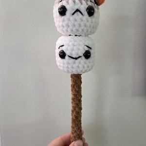 May include: A handmade crochet marshmallow on a stick. The marshmallow has two stacked white plush balls with black bead eyes and smiling faces. The top marshmallow has a brown and orange flame detail. The stick is brown.