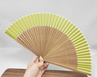 Könnte beinhalten: Ein offener Handfächer in Hellgrün und Beige. Der Fächer hat ein plissiertes Design mit einem hellgrünen Oberteil und beigefarbenen Holzrippen. Die Holzrippen sind mit einem weißen Punktmuster verziert. Der Fächer wird von einer Person gehalten.