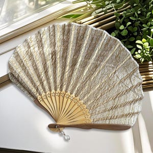 May include: A decorative hand fan with a light beige and gold patterned fabric. The fan's wooden handle and ribs are a light brown colour, with a decorative crystal charm hanging from the base. The fan is open, displaying its full shape.