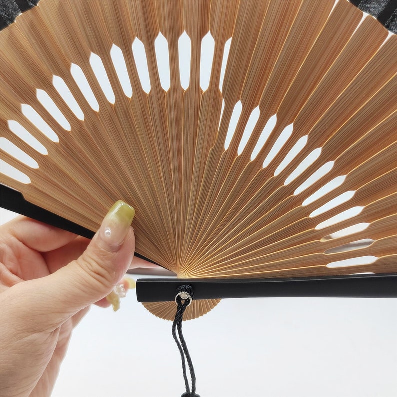 Puede incluir: Un abanico japon&eacute;s tradicional con marco de madera y tela negra. El abanico est&aacute; abierto y tiene un mango negro con una cuerda atada.