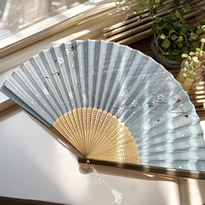 Op de afbeelding: Een lichtblauwe vouwwaaier met een houten handvat en delicate witte bloemenmotieven. De waaier is open en heeft een lichtblauwe zijden stof met een subtiele glans.