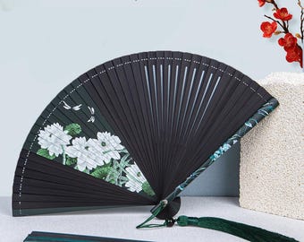 May include: A black folding fan with a white floral design and a green tassel. The fan is made of bamboo and has a delicate, intricate design.