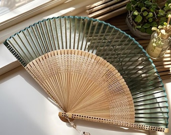 May include: A partially open hand fan with a bamboo frame and a translucent green fabric. The fan has a decorative beaded tassel and is resting on a white surface. The fan is a traditional accessory.