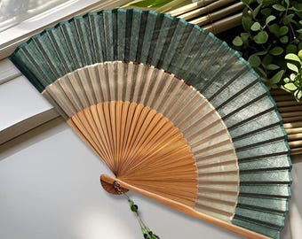 May include: A hand fan with a wooden handle and a pleated fabric design. The fan features a gradient of colors, transitioning from a light beige to a deep green. A decorative green bead tassel hangs from the handle.