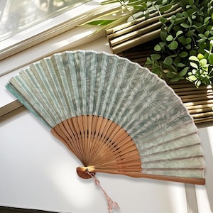 May include: A folding hand fan featuring a light green and white floral pattern on a pleated fabric surface. The fan has a light brown wooden frame with thin, vertical slats and a small, dangling pink beaded charm attached to the base.