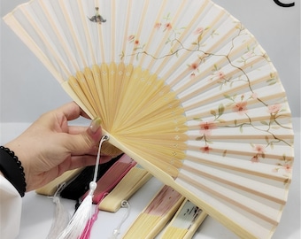 May include: A white folding fan with a floral design and a bamboo handle. The fan is decorated with pink flowers and green leaves. The fan is closed and has a tassel.