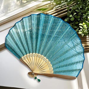 May include: A turquoise hand fan with a scalloped edge and a wooden handle. The fan's fabric has a textured appearance and is accented with a dark blue trim. The fan is decorated with a small green jewel.