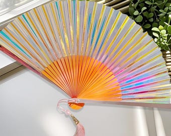 Può includere: Un ventaglio a mano aperto e vibrante con una sfumatura di colori. Il ventaglio presenta una base arancione brillante che sfuma in strisce gialle, blu e rosa. Un nappa rosa pende dalla base, aggiungendo un tocco decorativo.