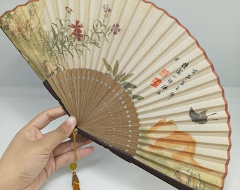 Puede incluir: Un abanico plegable beige con un mango de madera marrón y una borla dorada. El abanico está decorado con un diseño floral y un pájaro en vuelo. El abanico tiene caracteres chinos escritos en él.