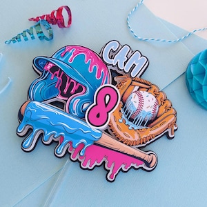 May include: A baseball-themed cake topper with a blue helmet, baseball bat, and glove, all with a dripping paint effect. The number 8 is in pink, and the name "CAM" is in blue. Additional party decorations are in the background.