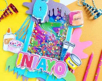May include: A colorful cake topper with the word "NIAYO" in rainbow letters, a blue number six, and a slime theme. The topper features a clear plastic container filled with colorful confetti and glitter, and the words "Slime" and "Glue" are visible on the topper.