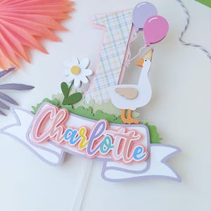 May include: A cake topper featuring a white goose with a pink hat holding two balloons, one purple and one pink. The goose is standing on a green grass base with a white banner that says "Charlotte" in rainbow colors. The number one is behind the goose and is decorated with a plaid pattern.