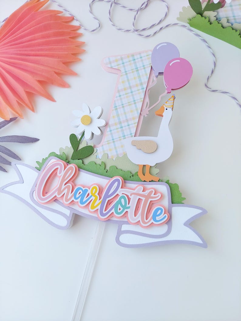 May include: A cake topper featuring a white goose with a pink hat holding two balloons, one purple and one pink. The goose is standing on a green grass base with a white banner that says "Charlotte" in rainbow colors. The number one is behind the goose and is decorated with a plaid pattern.