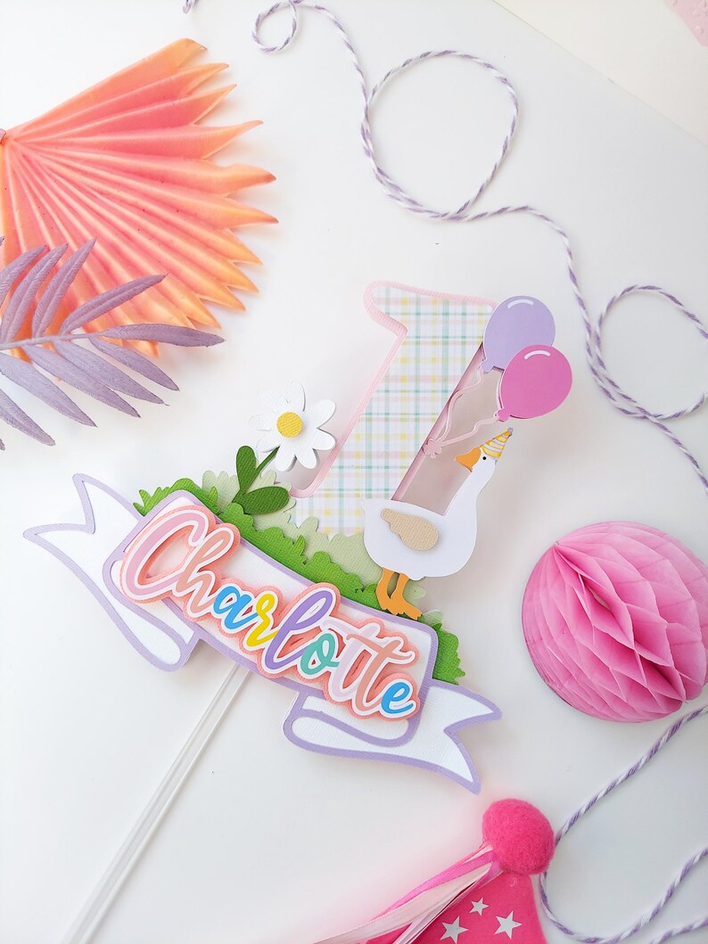 May include: A pastel-colored cake topper featuring a goose with a party hat, balloons, a daisy, and a banner that says "Charlotte".