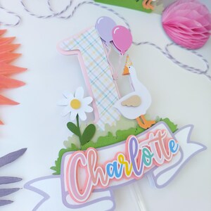 May include: A cake topper featuring a pink and yellow plaid number one, a white goose with a party hat, a white daisy, green grass, and a white banner with the name "Charlotte" in rainbow colors.