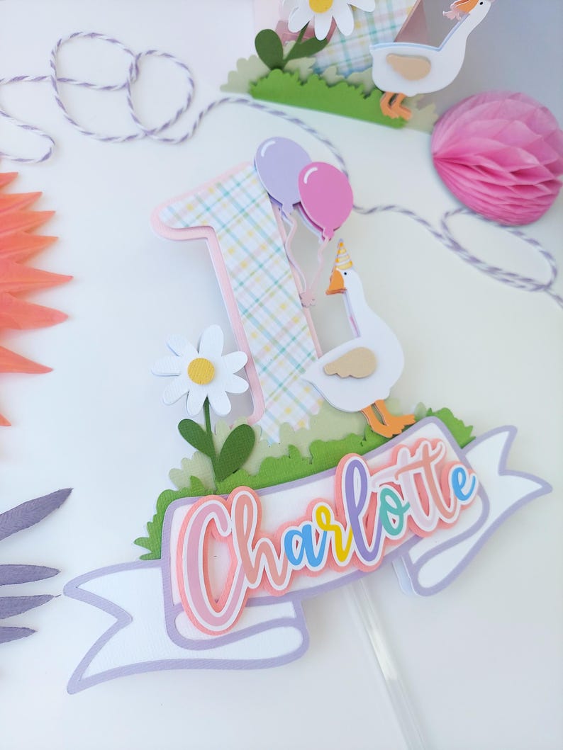 May include: A cake topper featuring a pink and yellow plaid number one, a white goose with a party hat, a white daisy, green grass, and a white banner with the name "Charlotte" in rainbow colors.