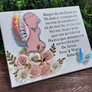 May include: A white canvas with a pink angel silhouette, gold glitter wings, and a blue butterfly. The canvas is decorated with paper flowers in shades of pink and white. The text on the canvas reads: "Angel de mi Guarda Mi dulce compañia. no me desampares ni de noche, ni de día. No me dejes sola. que me perdería Hasta que amanezca en los brazos De Jesus Jose y Maria Amen"