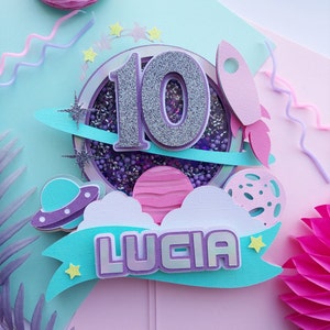 May include: A purple and teal paper cake topper with a sparkly silver number 10, a rocket, planets, and the name "Lucia".
