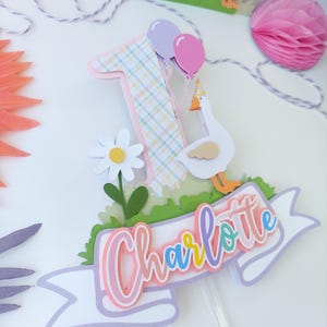 May include: A pink, white, and green cake topper with a number one, a goose, a daisy, and a banner that says "Charlotte".
