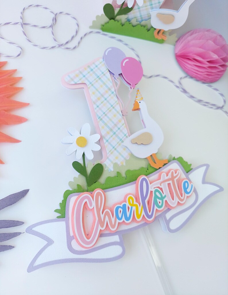 May include: A pink, white, and green cake topper with a number one, a goose, a daisy, and a banner that says "Charlotte".