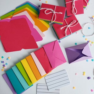 May include: Assortment of colorful envelopes in various sizes and shapes, including a set of rainbow envelopes, a stack of red envelopes, and a single purple envelope. The envelopes are arranged on a white surface with scattered heart-shaped confetti.
