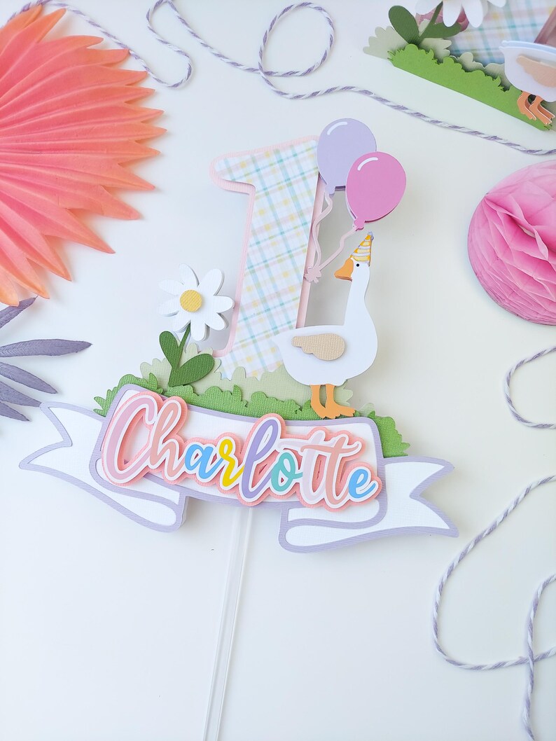 May include: A pink, white, and yellow plaid number one cake topper with a white goose holding pink and purple balloons. The topper also includes a white banner with the name "Charlotte" written in rainbow colors.