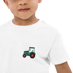 Puede incluir: Una camiseta blanca con un tractor verde bordado en la parte delantera.